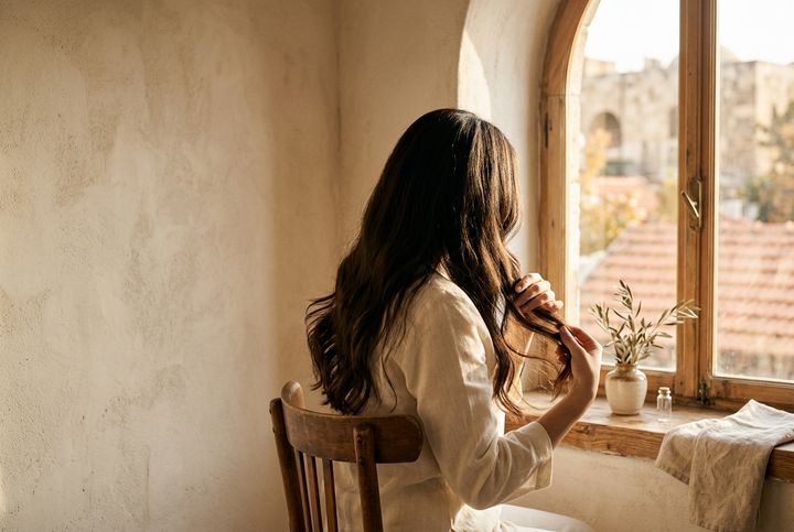 مشهد تعبيري لامرأة ترتّب أطراف شعرها قرب نافذة مضيئة